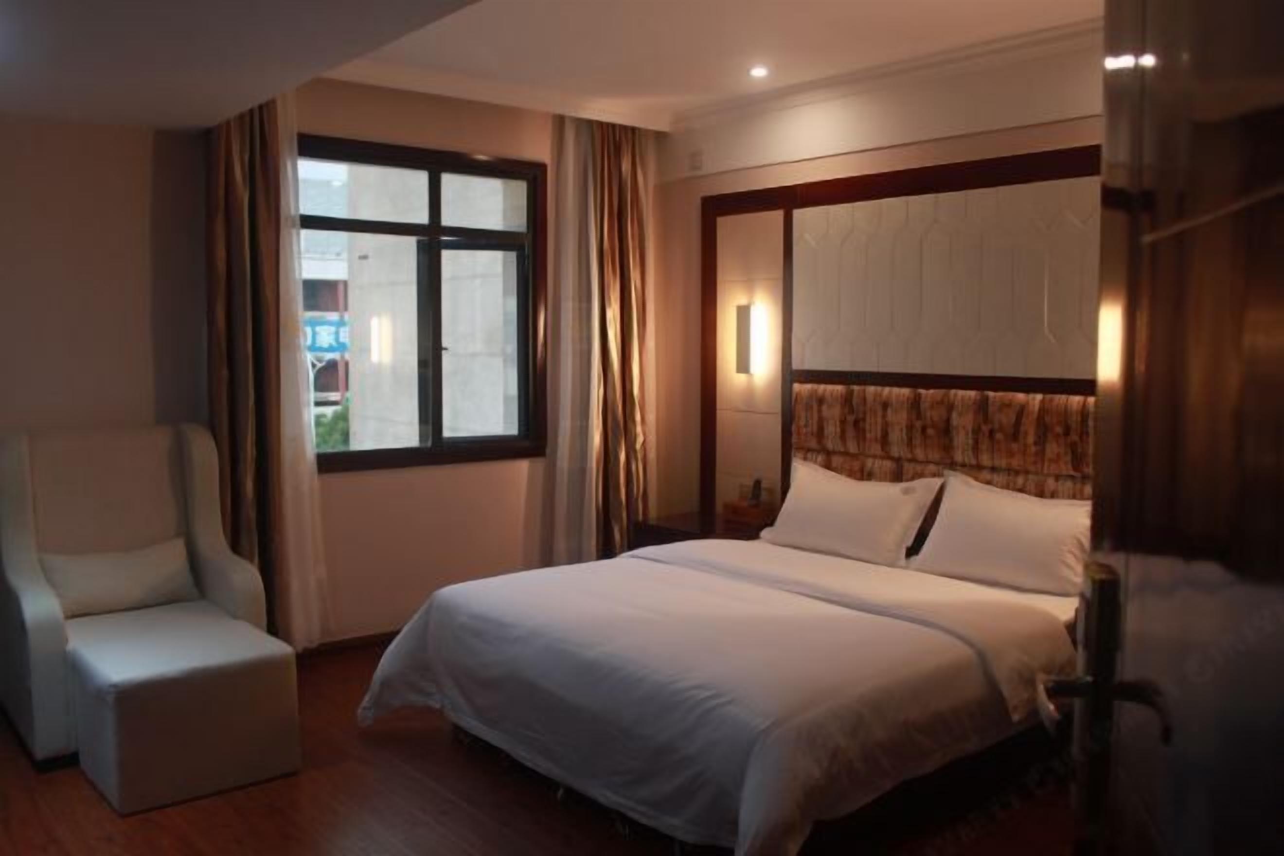A hotel room with a bed, a chair, a window, and a wall-mounted light fixture.