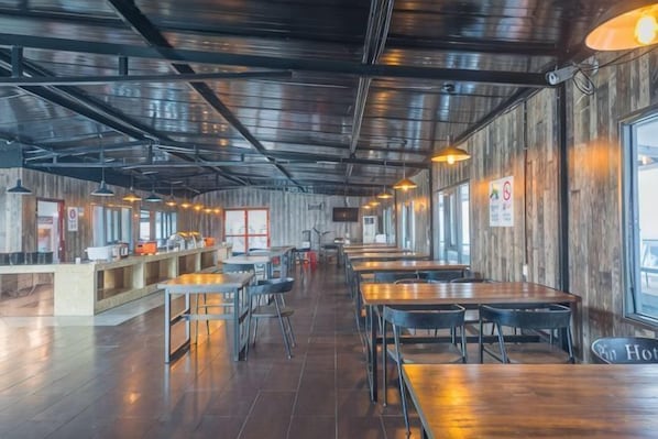 A restaurant with long communal tables, a counter with food items, and large windows along the walls.