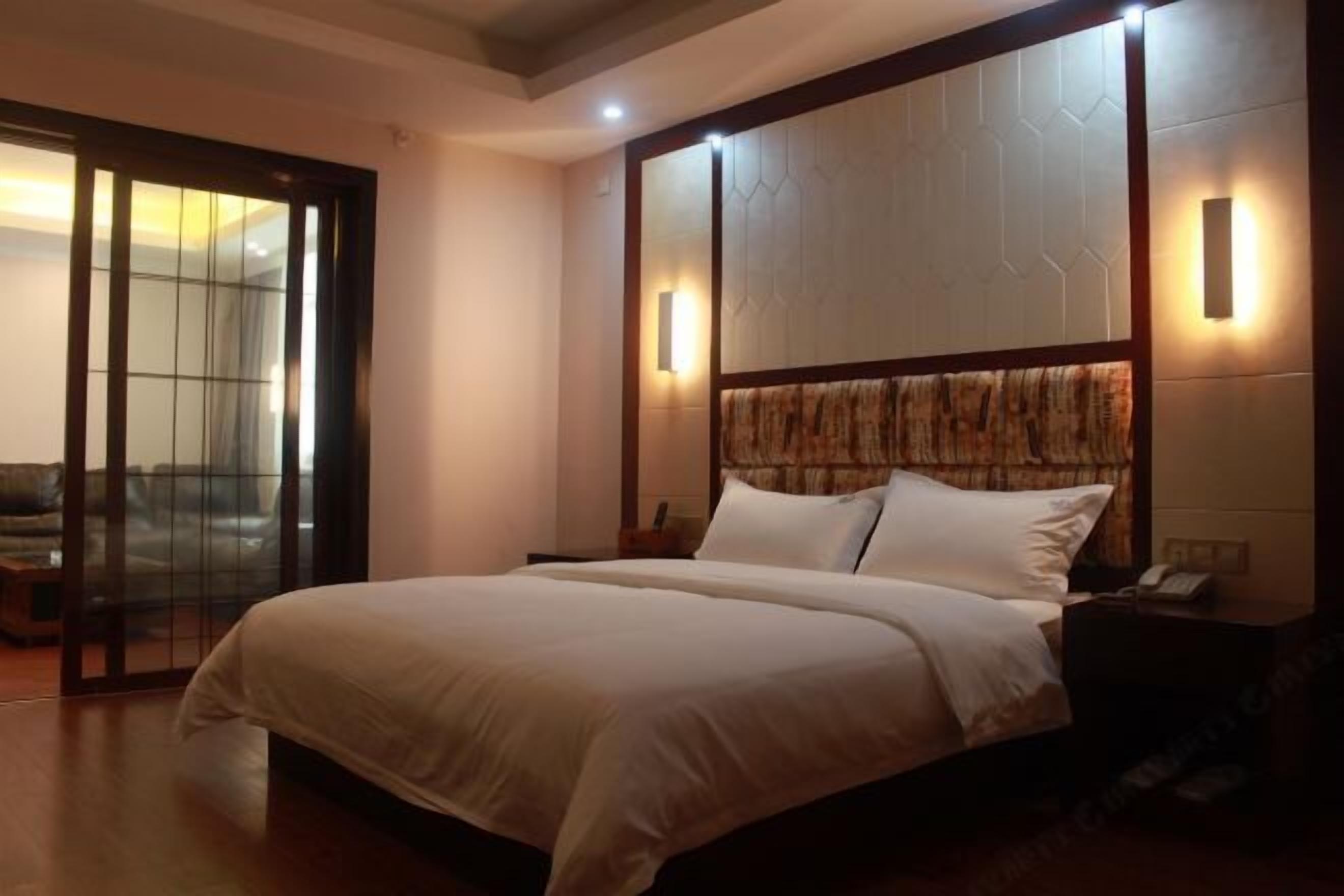 A hotel room with a large bed, a wooden headboard, a nightstand, and a sliding glass door leading to a balcony.