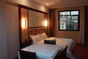 A hotel room with a bed, a bedside table, a chair, a window with a view of buildings, and a wall-mounted mirror.