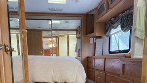 A compact RV interior with a bed, kitchenette, and a small window.