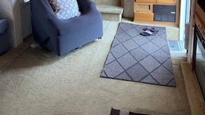 A room with a ceiling fan, a blue armchair, a patterned rug, wooden cabinets, and a door leading to another room.