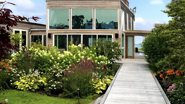 Una casa moderna con una terraza de madera que conduce a un vidrio, rodeada de jardines coloridos.