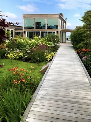 A modern house with a wooden deck leading to a glass door, surrounded by a variety of colorful gardens. - Horace Gifford Design - Modernist Gem on Fire Island - Pines (Fire Island)