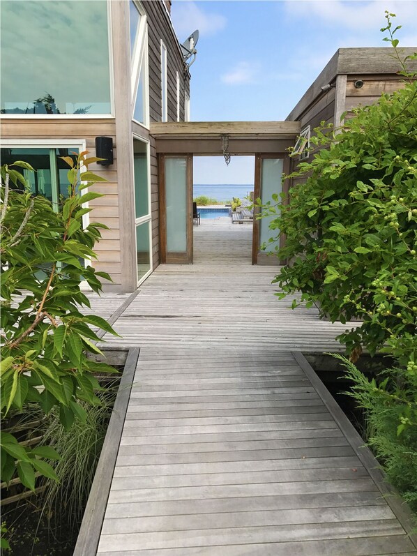 A wooden deck leading to a building with a glass door, a satellite dish, and a view of the ocean. - Horace Gifford Design - Modernist Gem on Fire Island - Pines (Fire Island)