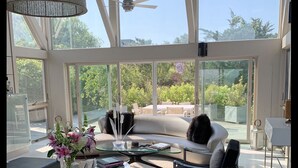 Un amplio salón con un gran ventanal que da a un patio exterior, una lámpara moderna y un ventilador de techo.
