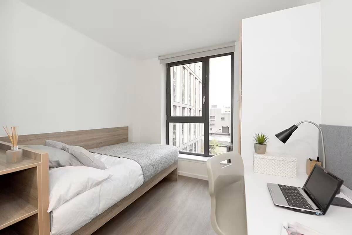 A modern bedroom with a bed, a desk with a laptop, a chair, a window with a view of buildings, and a small plant on the desk.