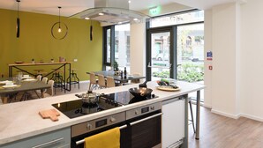 A modern kitchen with a central island, dining area, and large windows offering a view of the street. - For Students Only Bright Ensuite at St Mungo's in Glasgow (Glasgow)