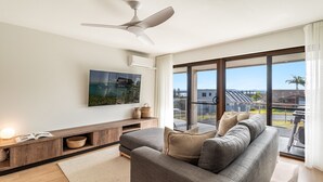 Un salon moderne avec un canapé gris, un ventilateur de plafond, une grande fenêtre donnant sur des bâtiments et des espaces verts, et une œuvre d’art accrochée au mur.