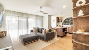Un salon moderne avec un canapé d’angle gris, une table basse en bois et un coin cuisine doté d’un crédence à motifs et de meubles de rangement en bois.