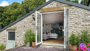 A stone house with a sloped roof, a sliding glass door leading to a living room with a sofa and a fireplace, surrounded by potted plants and flowers. - 1 bedroom accommodation in Buxton (Buxton)