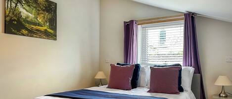 A bedroom with a bed, a window with curtains, a nightstand, a lamp, and a painting on the wall.