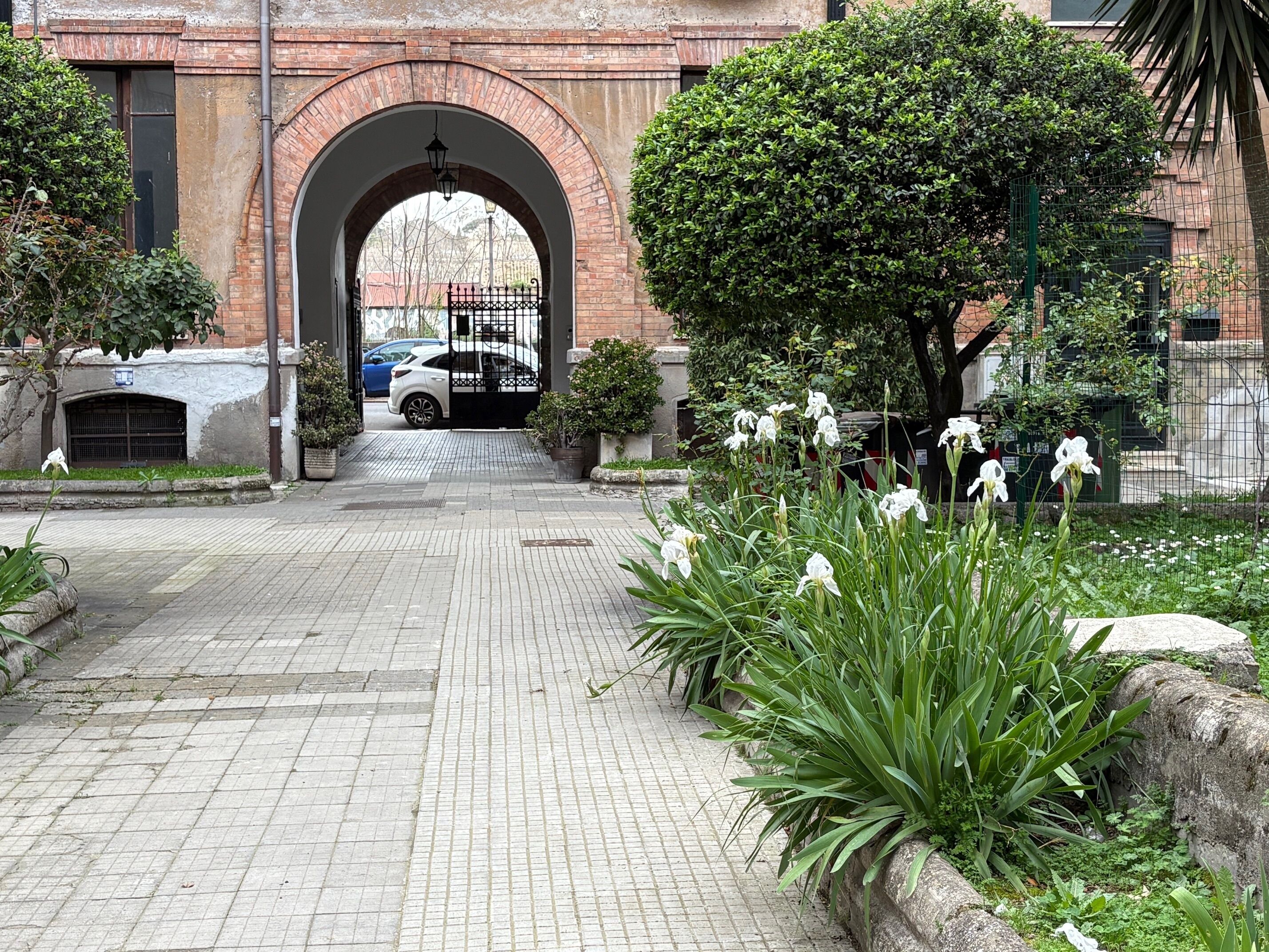 A paved pathway leading to a brick archway, surrounded by greenery and white flowers.