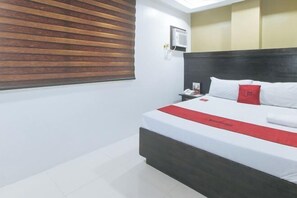 A hotel room with a bed, a nightstand, a red and white bedspread, a wooden slat window blind, and a wall-mounted air conditioning unit. - RedDoorz @ JQV HOTEL Camarin Caloocan (Caloocan)