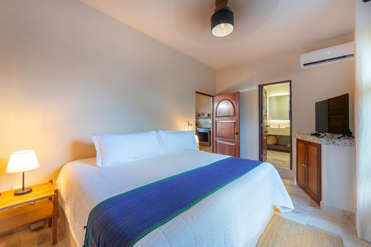 A hotel room with a large bed, a bedside table with a lamp, a wooden wardrobe, and an air conditioning unit.
