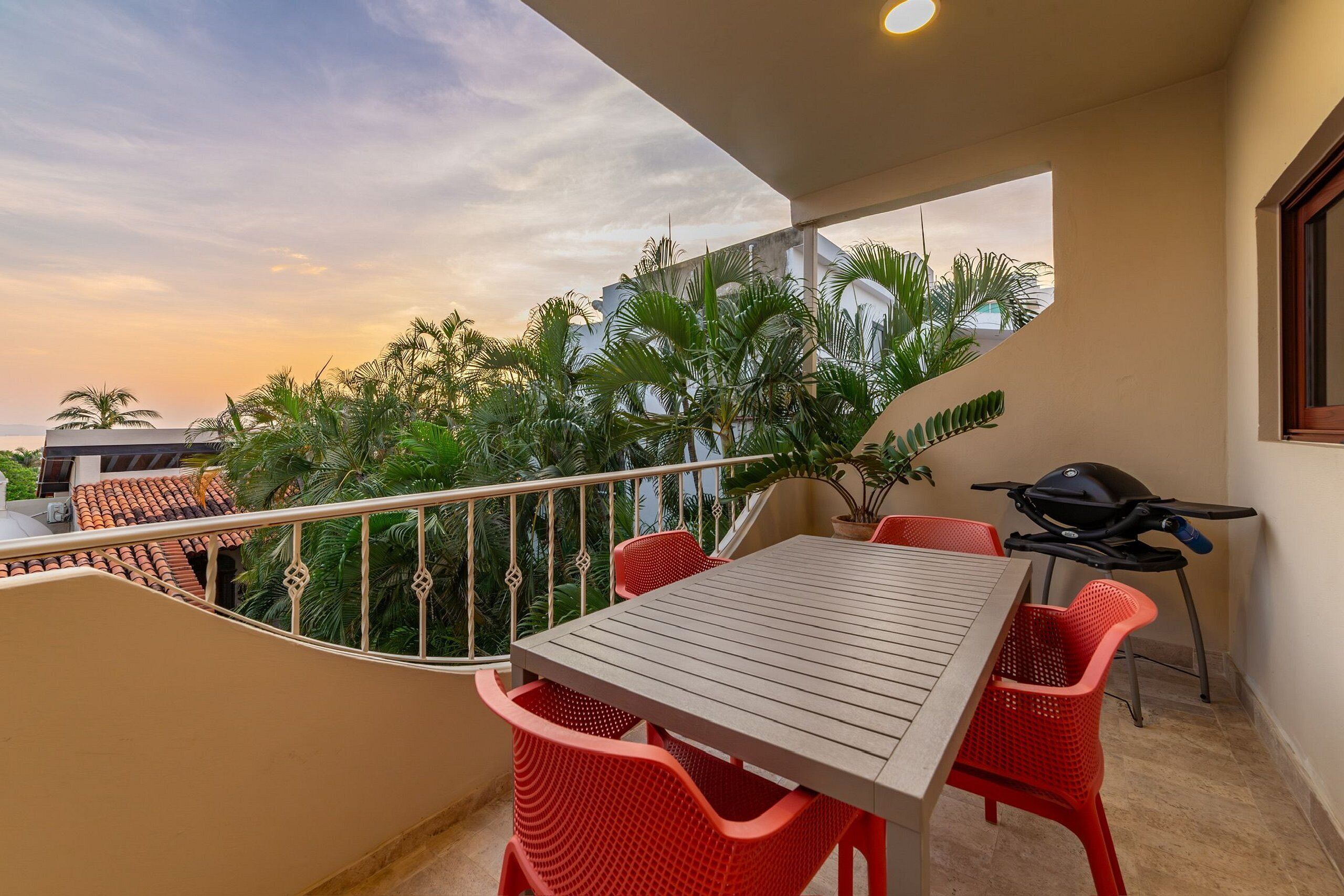 A balcony with a table and chairs, a grill, and a view of palm trees and buildings.