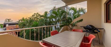 A balcony with a table and chairs, a grill, and a view of palm trees and buildings.