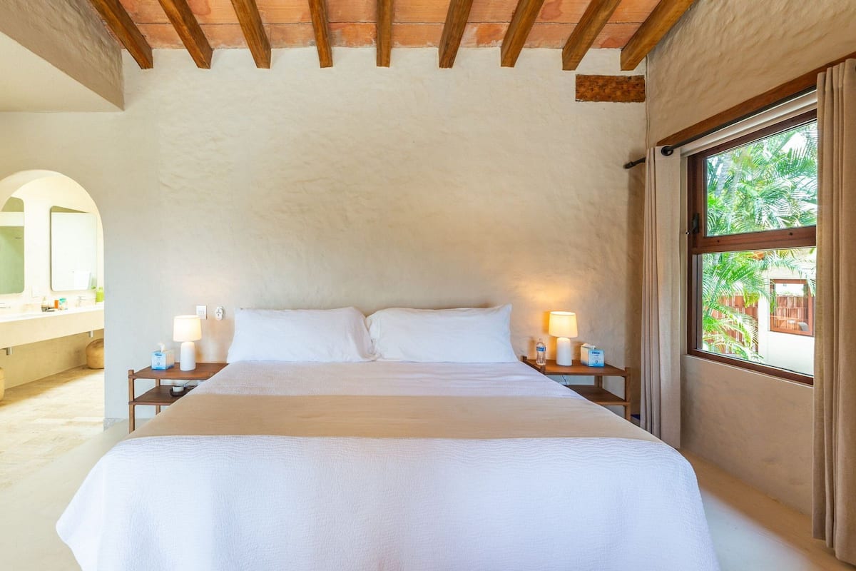 A bedroom with a large bed, wooden headboard, two bedside tables with lamps, a window with curtains, and a view of greenery outside.