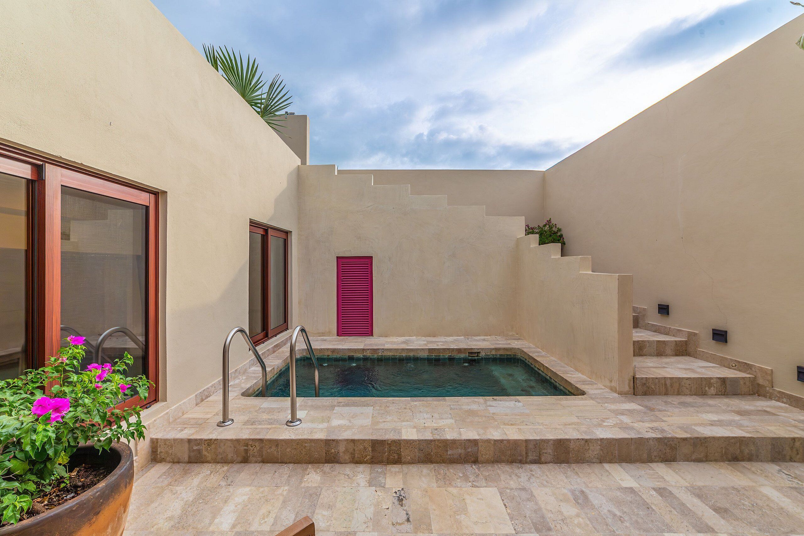 A swimming pool with steps leading up to it, surrounded by beige walls and a potted plant with pink flowers.