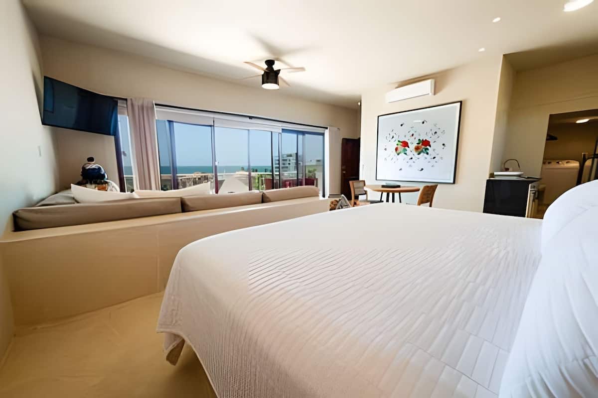 A hotel room with a large bed, a seating area with a view of the ocean, a ceiling fan, and a dining area.
