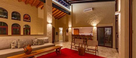A spacious living area with a high ceiling, a stone wall, a wooden bench, a red rug, and a seating area with cushions.