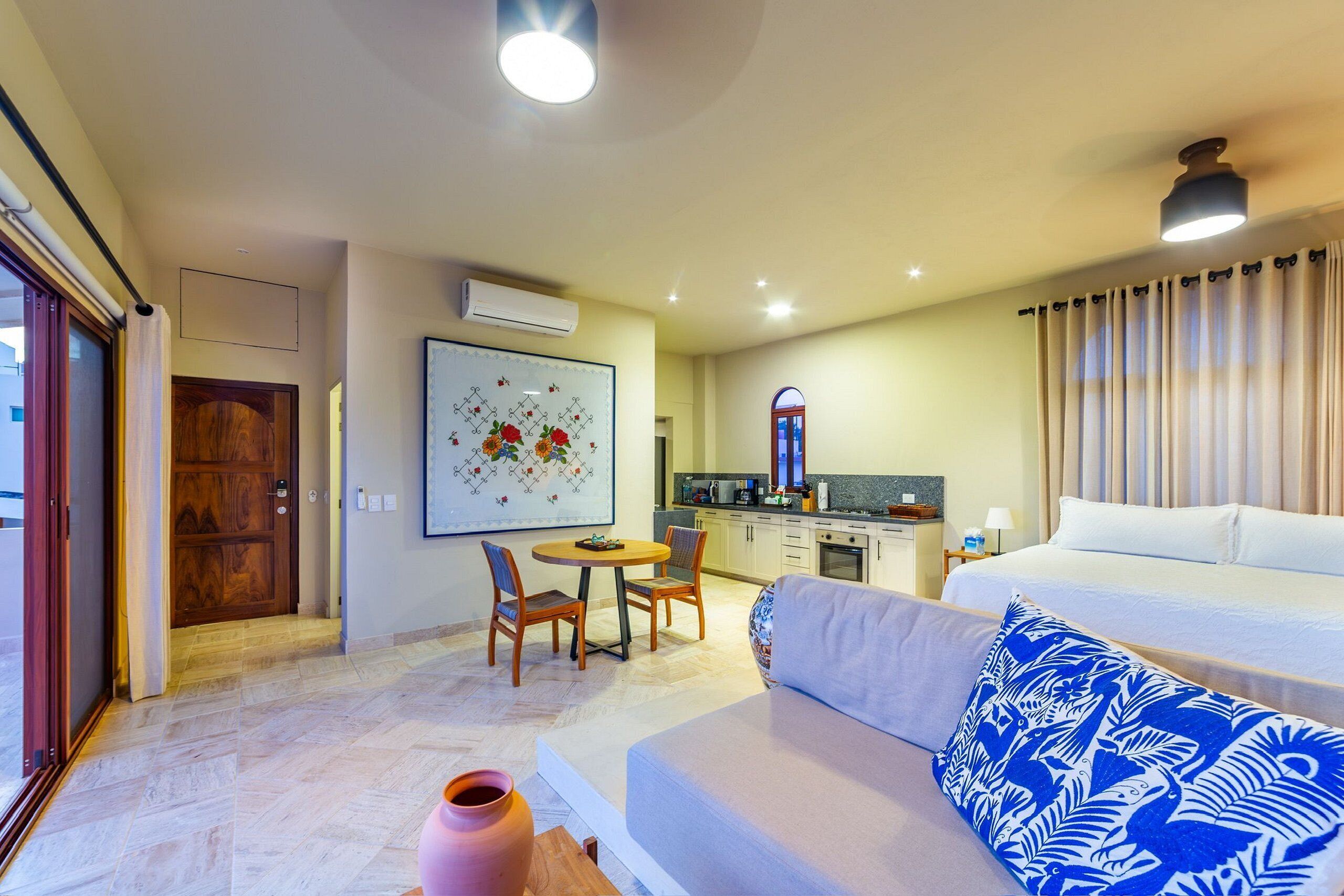 Habitación de hotel con cama, sofá, mesa de comedor con sillas, zona de cocina y un ventanal con cortinas.
