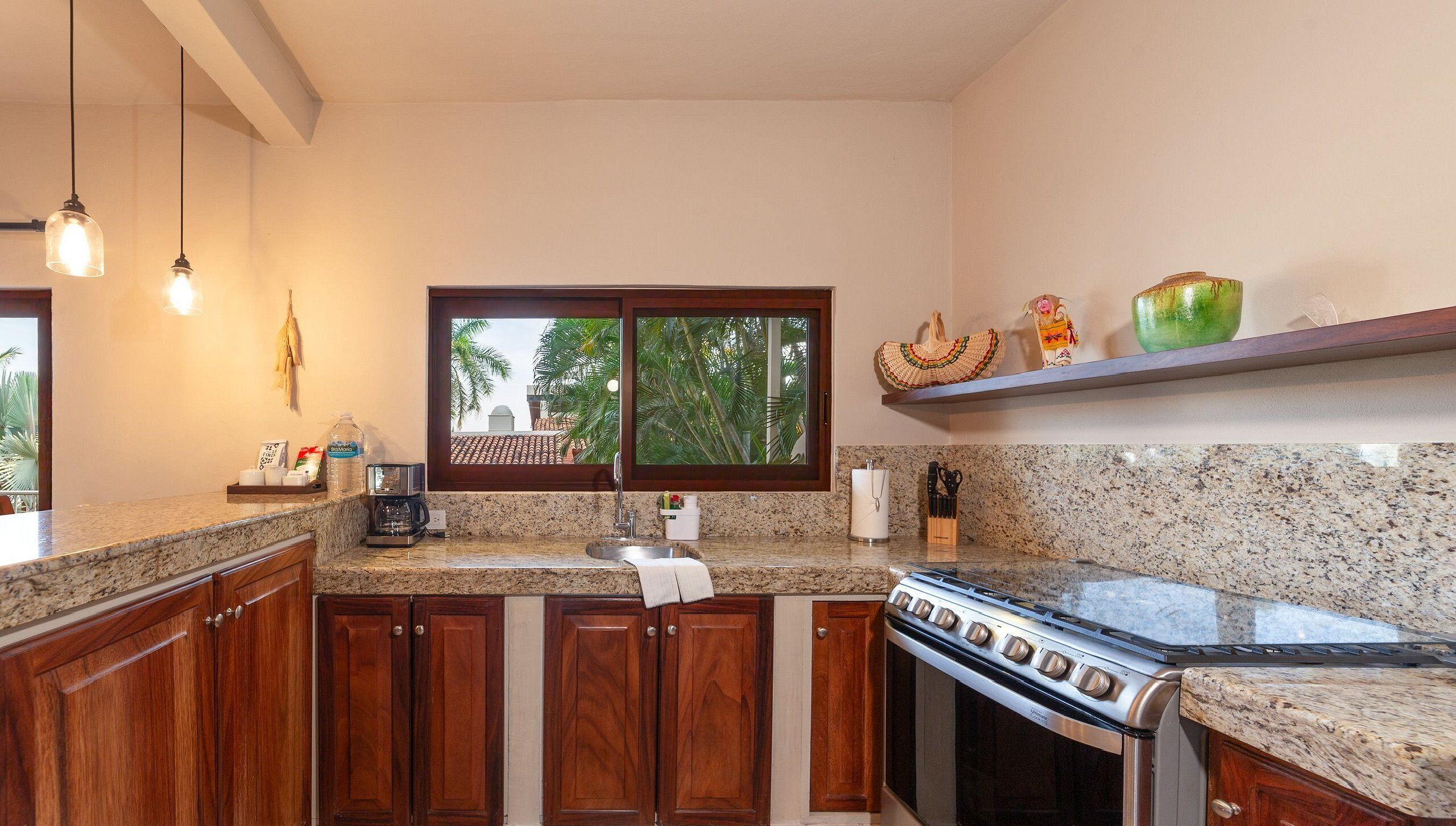 A kitchen with wooden cabinets, a granite countertop, a stove, a sink, and a window with a view of palm trees.