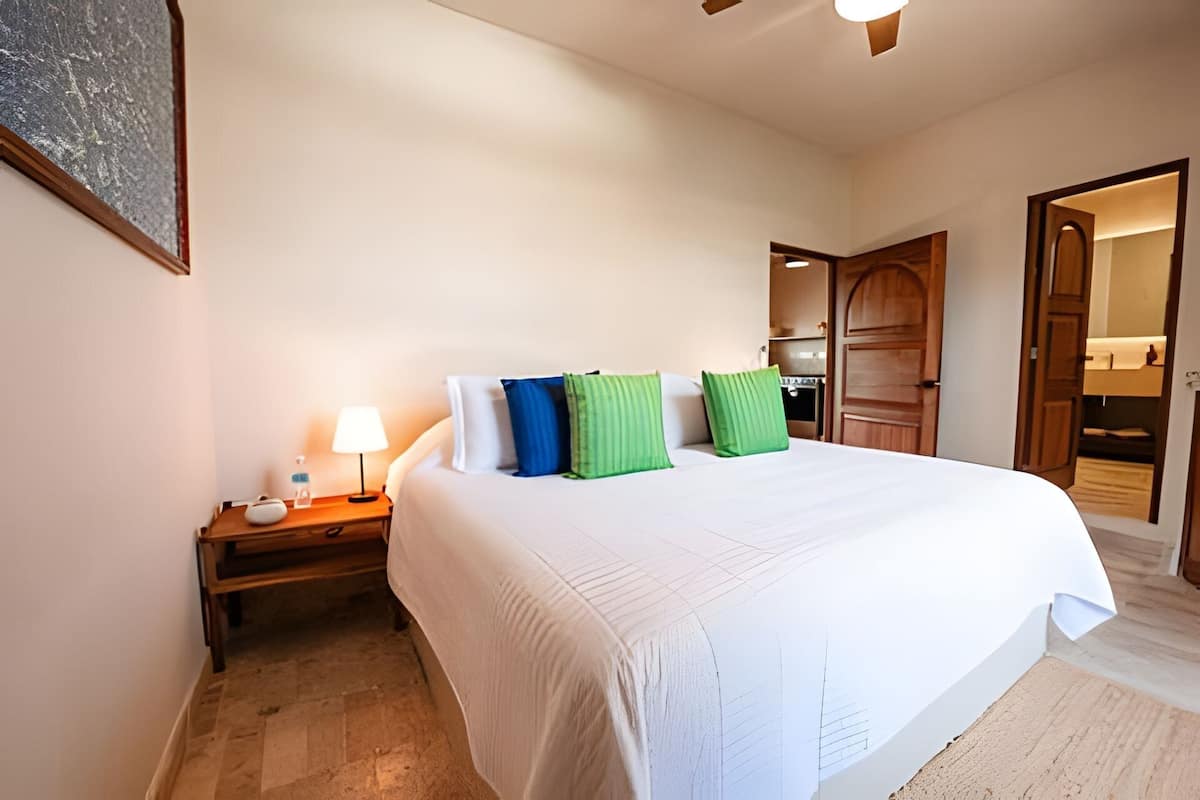 A bedroom with a large bed, a nightstand with a lamp, and a wooden door.