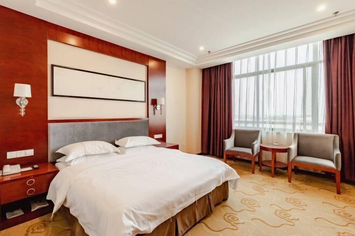 Une chambre d’hôtel avec un grand lit, deux tables de chevet, un bureau, deux chaises et une grande fenêtre avec des rideaux.