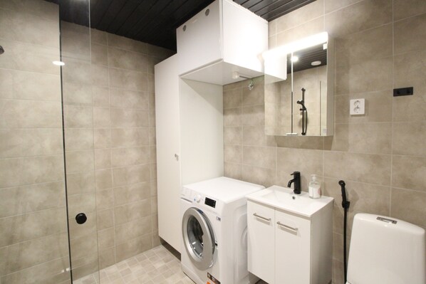 Une salle de bain moderne dotée d’une douche à parois de verre, d’un lave-linge encastré et d’un lavabo blanc avec un distributeur de savon.