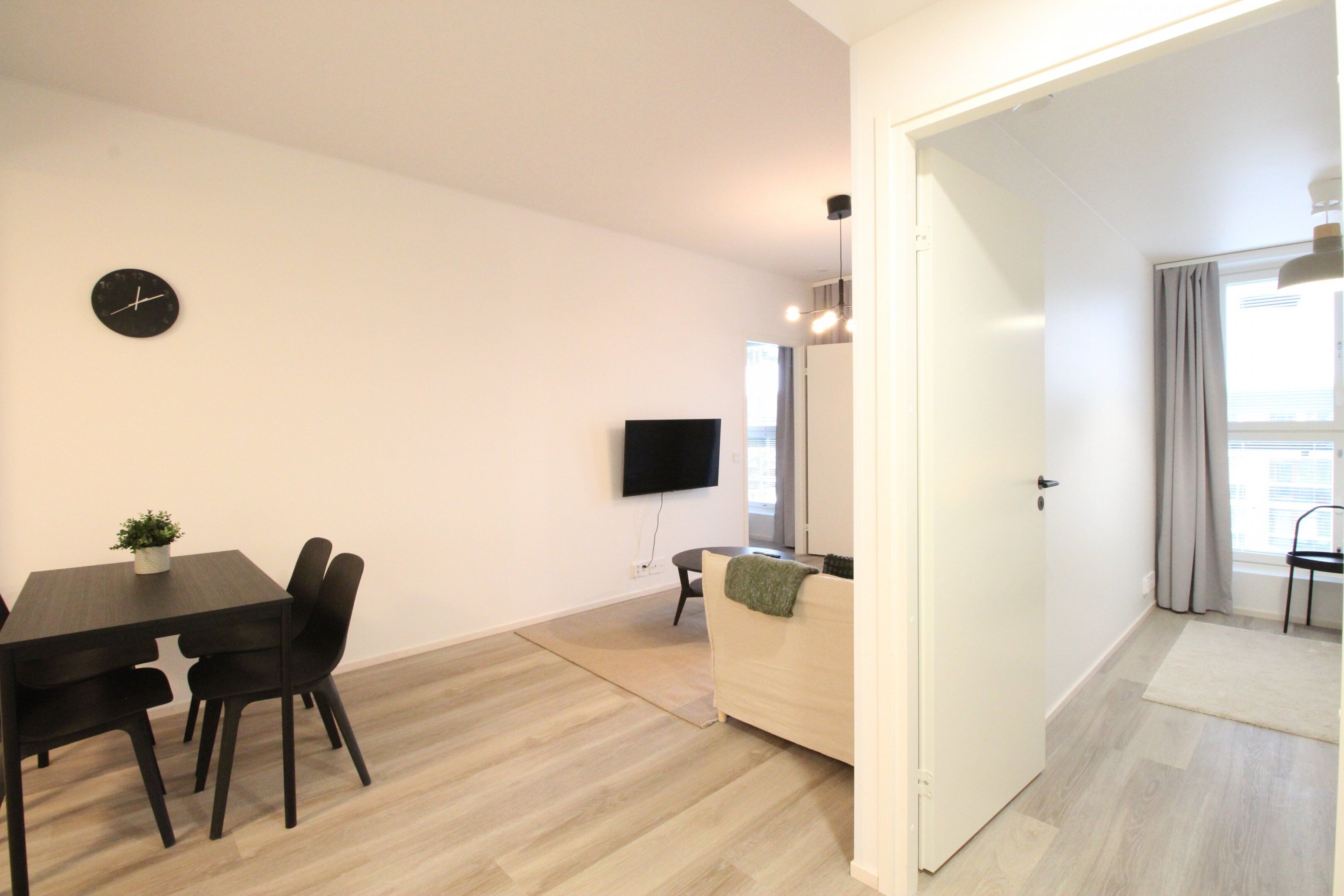 Una sala moderna con una mesa de comedor, sillas, un sofá, un televisor montado en la pared y una puerta que da a otra habitación.