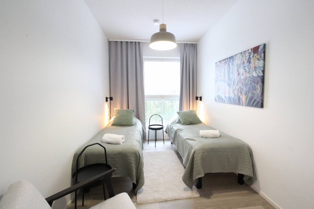 Una habitación con dos camas, una silla, una mesita, un cuadro en la pared y una ventana con cortinas.