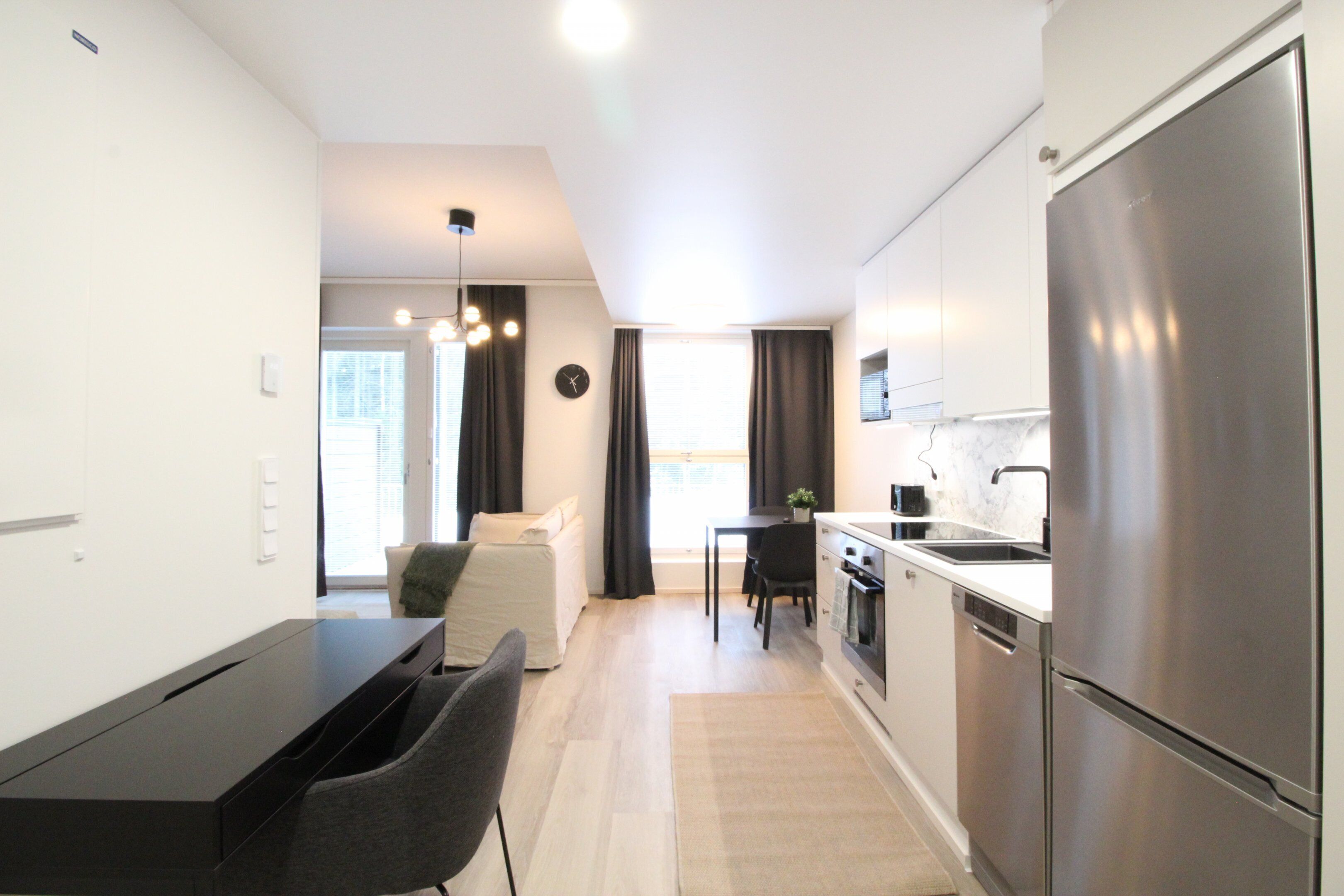 A modern kitchen with a stainless steel refrigerator, a dining area with a table and chairs, and a sofa in the background.