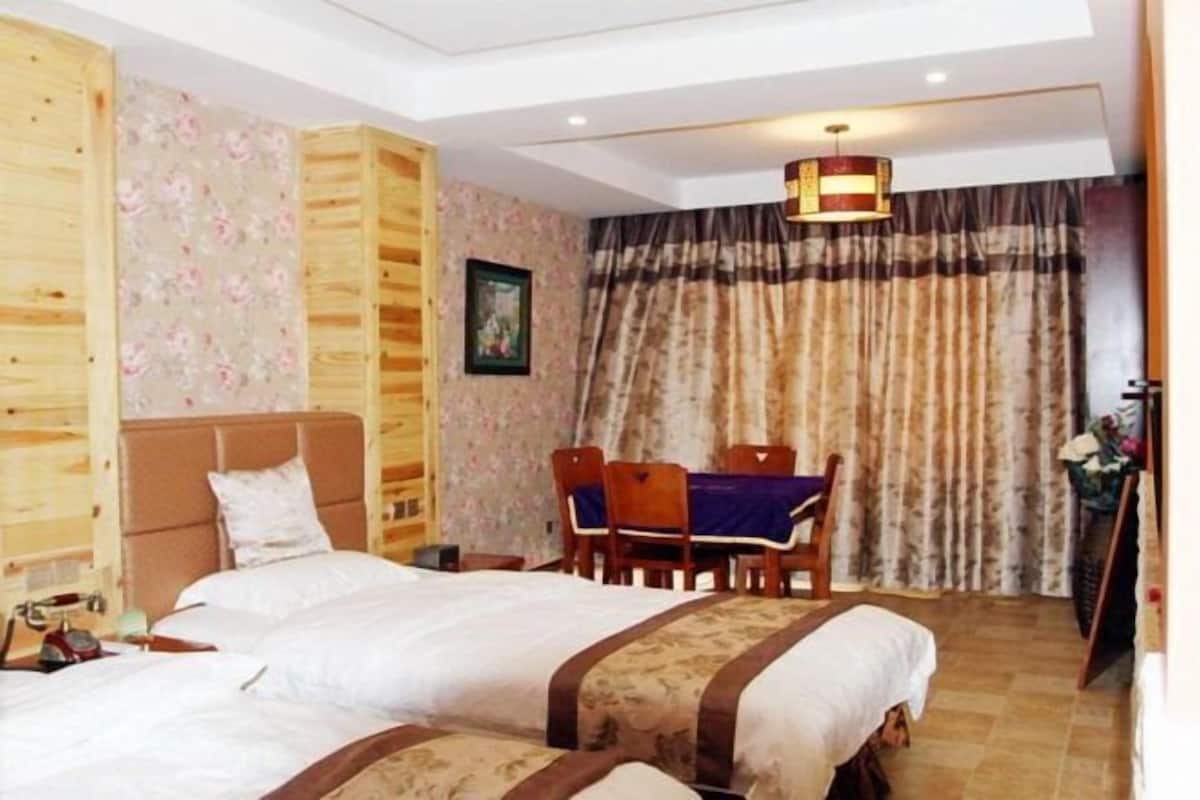 A bedroom with two beds, a wooden headboard, a table with chairs, and a large window with curtains.