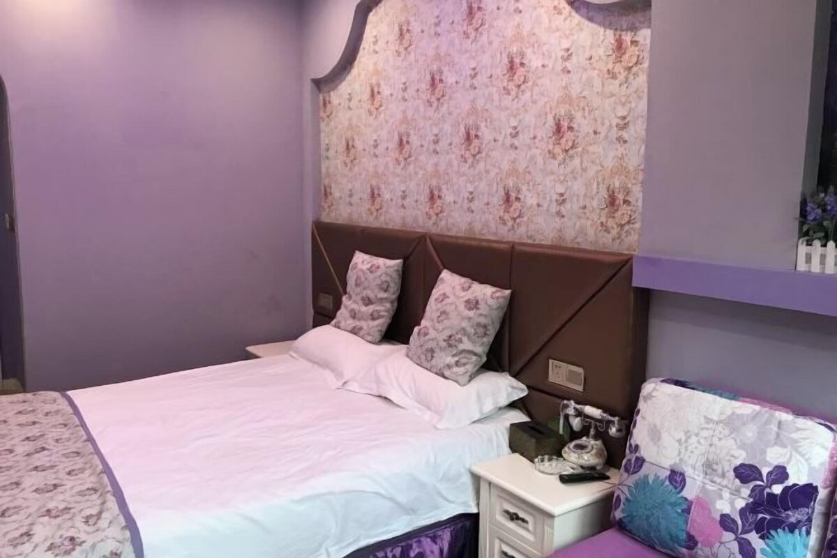 Ein ordentlich bezogenes Bett mit einem blumigen Überwurf, ein Kopfteil mit floralem Muster und ein Nachttisch mit Telefon und kleiner Lampe.