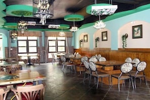 Un interior de restaurante con varias mesas y sillas, ventanas amplias y lámparas colgantes.