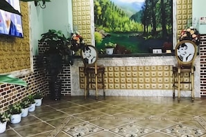 Un área de recepción con pared de ladrillo, piso de baldosas y un cuadro grande de un paisaje. Hay plantas en macetas y una silla de madera con un espejo.