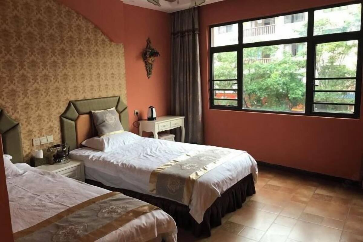 A hotel room with two beds, a window with a view of greenery, a nightstand with a lamp, and a wall-mounted decoration.