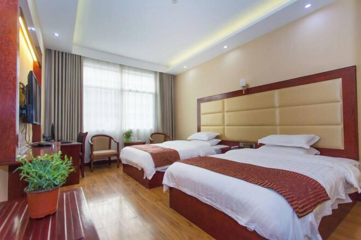 A hotel room with two beds, wooden flooring, a large window with curtains, a television on the wall, and a potted plant.