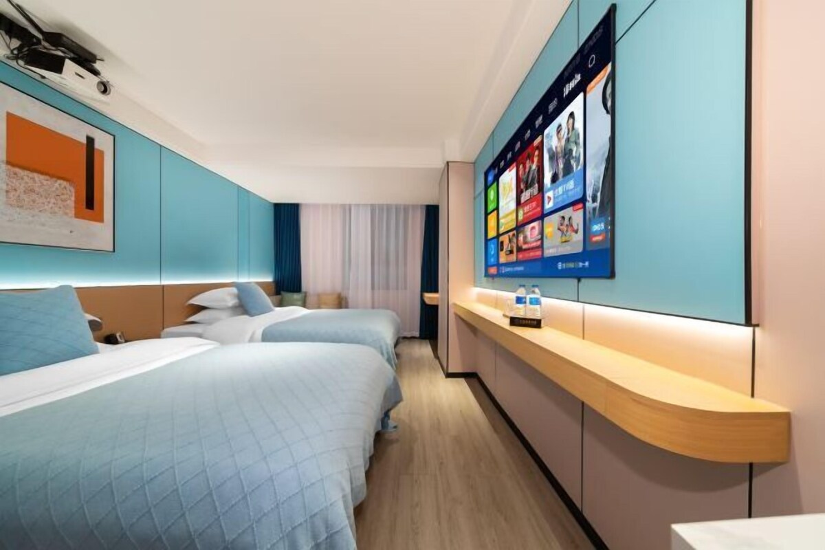 A hotel room with two beds, a bedside table, a large digital screen displaying content, and a window with curtains.