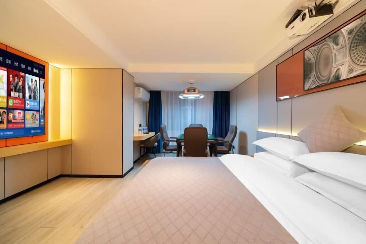 A hotel room with a bed, a desk with a chair, a television mounted on the wall, and a window with curtains.