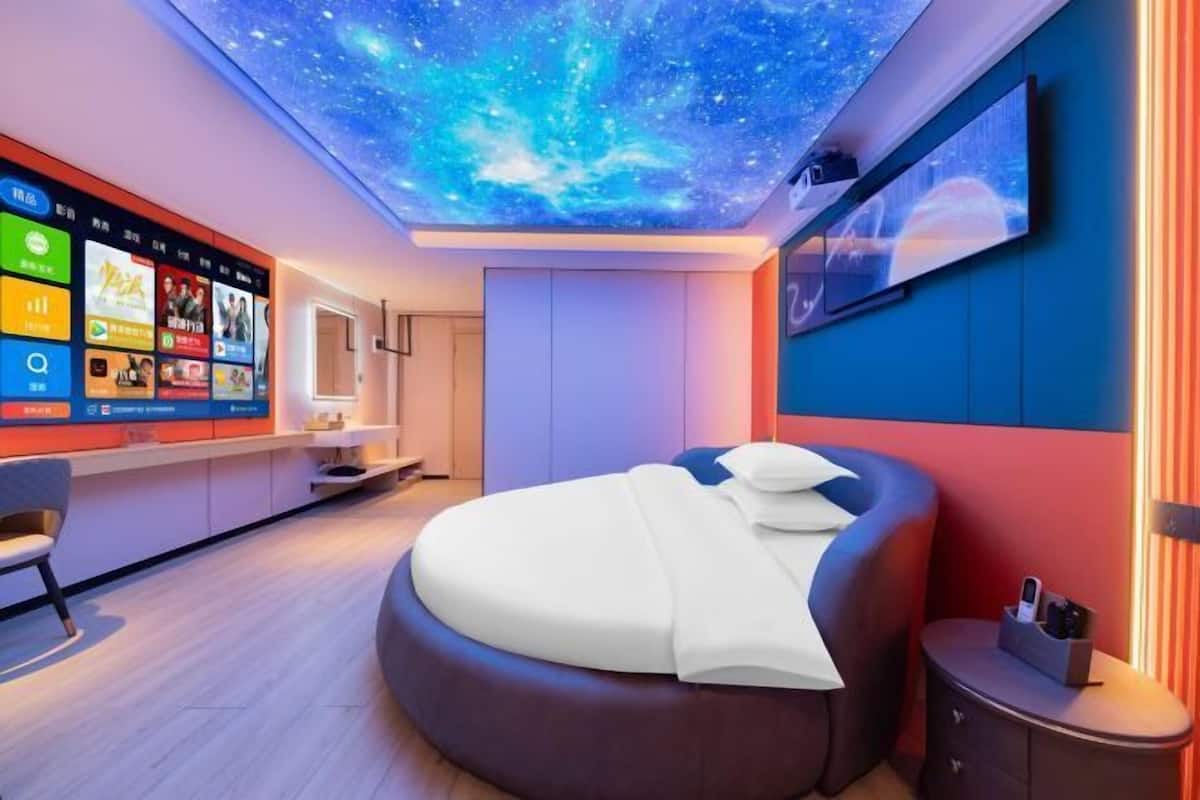 A modern bedroom with a curved bed, a digital sky ceiling, a TV on the wall, and a bedside table with a phone and charger.
