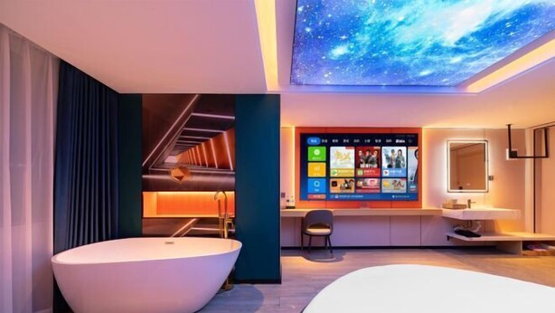 Une chambre d’hôtel moderne dotée d’un grand lit, d’une baignoire, d’un bureau avec un ordinateur et d’un téléviseur fixé au mur, qui affiche une interface aux couleurs vives.