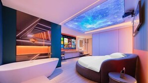 Une chambre d’hôtel moderne dotée d’un grand lit, d’un espace salon aux lignes courbes, d’un téléviseur intégré et d’un projecteur au plafond qui projette un ciel étoilé.