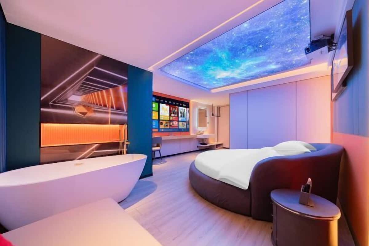 A modern hotel room with a curved bed, a large window, a ceiling projection, and a wall-mounted TV.