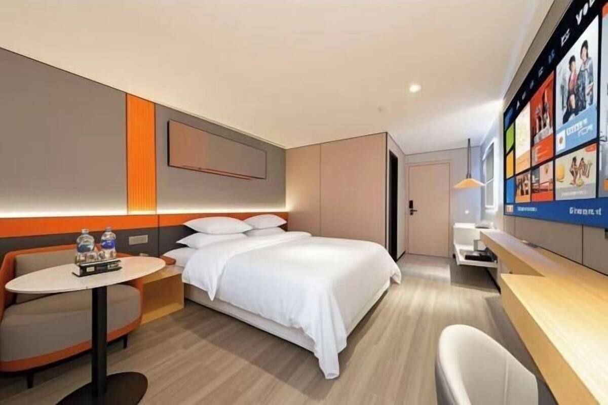 A modern hotel room with a bed, a desk, a chair, a TV mounted on the wall, and a small table with water bottles.
