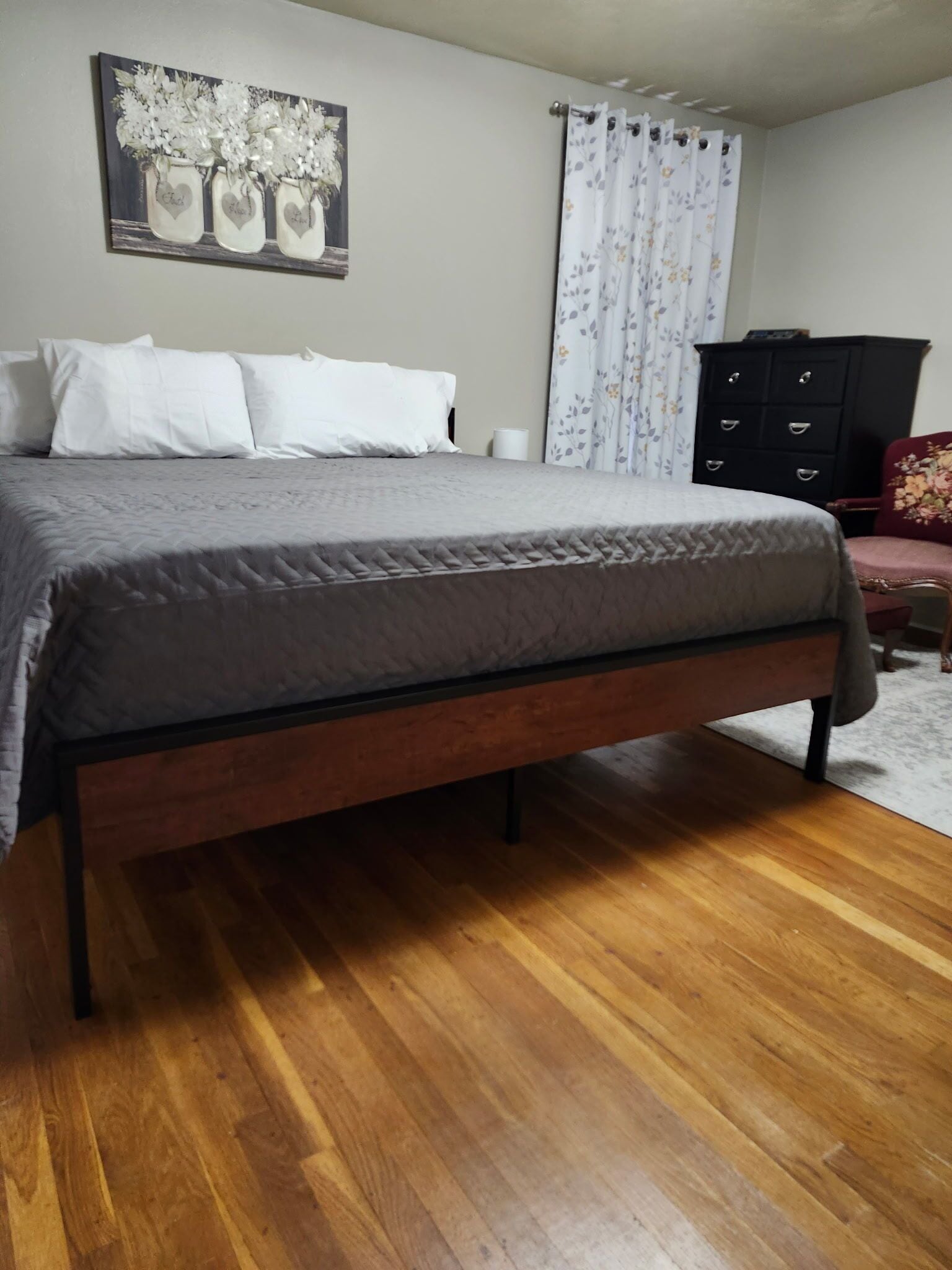 Una camera da letto con un letto, pavimento in legno, una cassettiera nera e una tenda floreale.