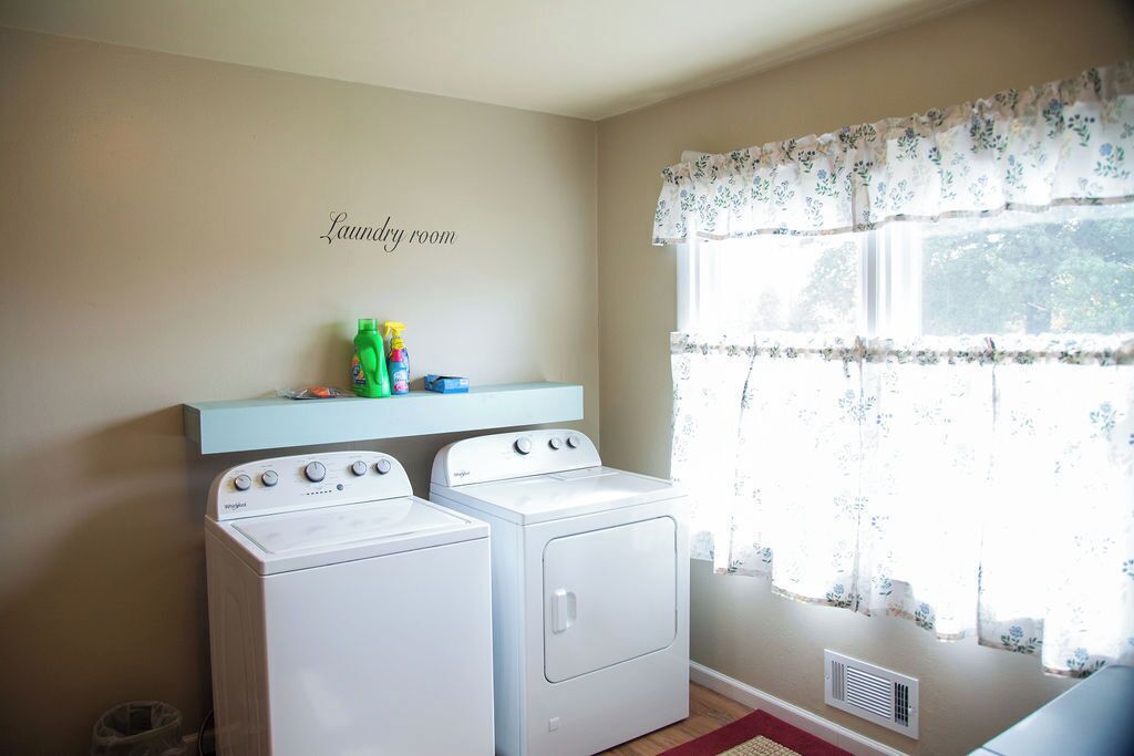 A laundry room with two white washing machines, a shelf with cleaning products, and a window with floral curtains.