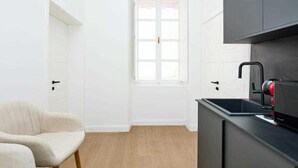 Eine kleine, helle Küche mit weißen Wänden, Holzboden, einem Fenster und einem Stuhl.