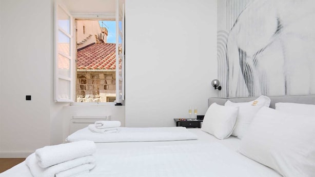 Una habitación de hotel con cama, mesita de noche, ventana con vistas a edificios y pared de mármol.